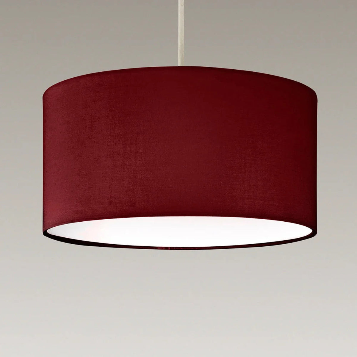 A large cylindrical drum pendant light shade in deep burgundy fabric, suspended by a thin white cord against a neutral light gray background.