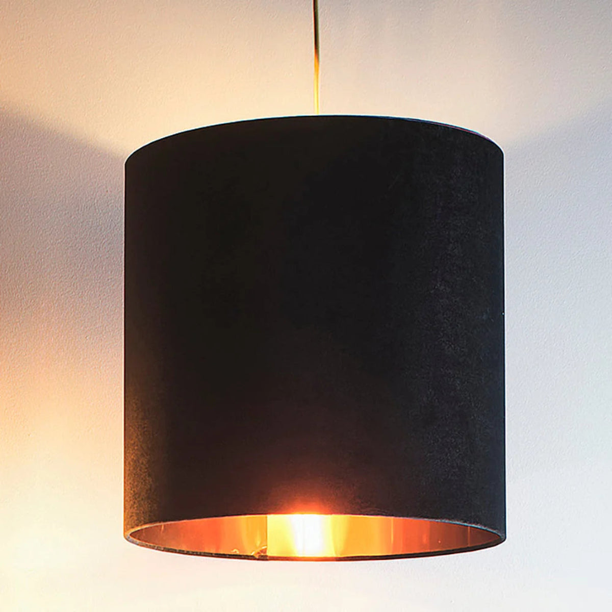 A black fabric drum pendant lampshade with a warm orange-gold light glowing from within, suspended by a thin cord against a plain wall.