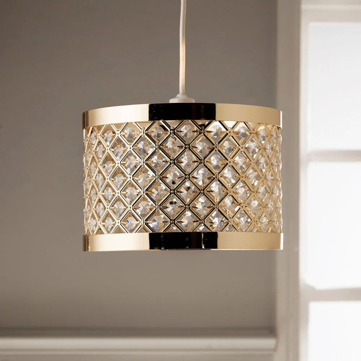 A modern drum pendant light with a gold-finished metal frame and a diamond-patterned clear crystal mesh shade, hanging against a neutral gray wall.