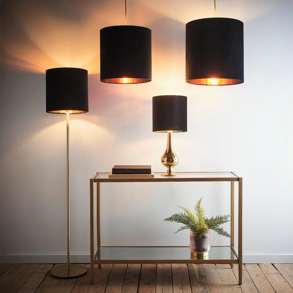 Three black fabric drum pendant lights in varying sizes hang against a white wall above a gold-framed glass console table. A matching black table lamp with a gold base sits on the table, and a floor lamp with a gold stand is on the left.