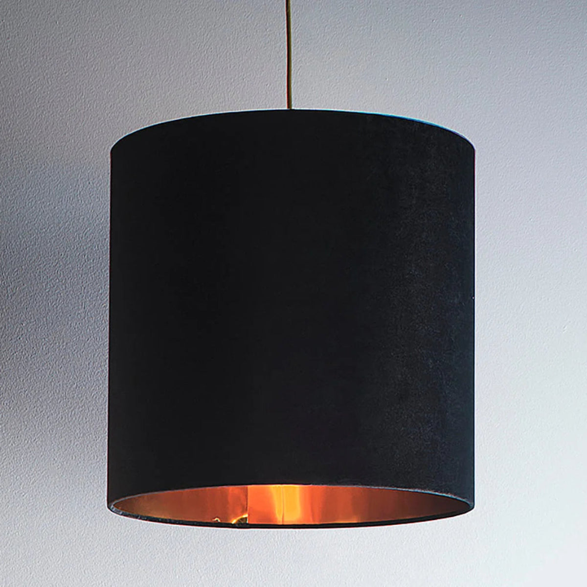 A black fabric drum pendant light shade with a warm copper-toned interior, shown hanging against a light gray wall.